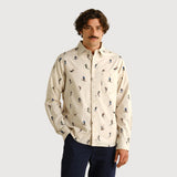 A man wearing a cream-colored long-sleeve button-down shirt with a repeating pattern of skiers, paired with navy blue trousers, standing against a plain light beige background.