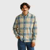 A man wearing a beige and blue plaid button-down shirt with dark blue jeans, standing against a solid light beige background.